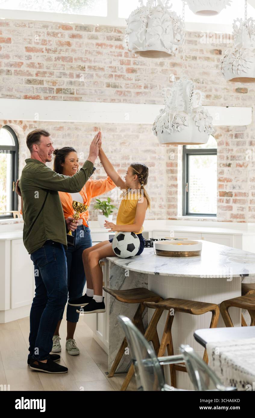 Padre e figlie festeggiano in cucina con pallone da calcio e trofeo. Famiglia, celebrazione, felicità, legame, bambini, sport Foto Stock