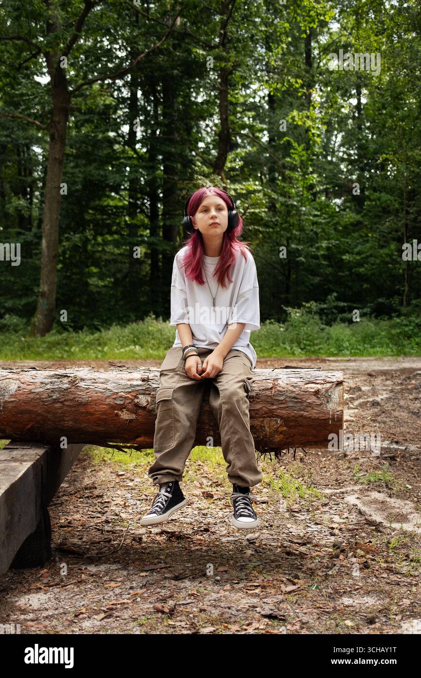 Adolescente con i capelli rosa che ascolta la musica in natura, seduto su un tronco d'albero Foto Stock