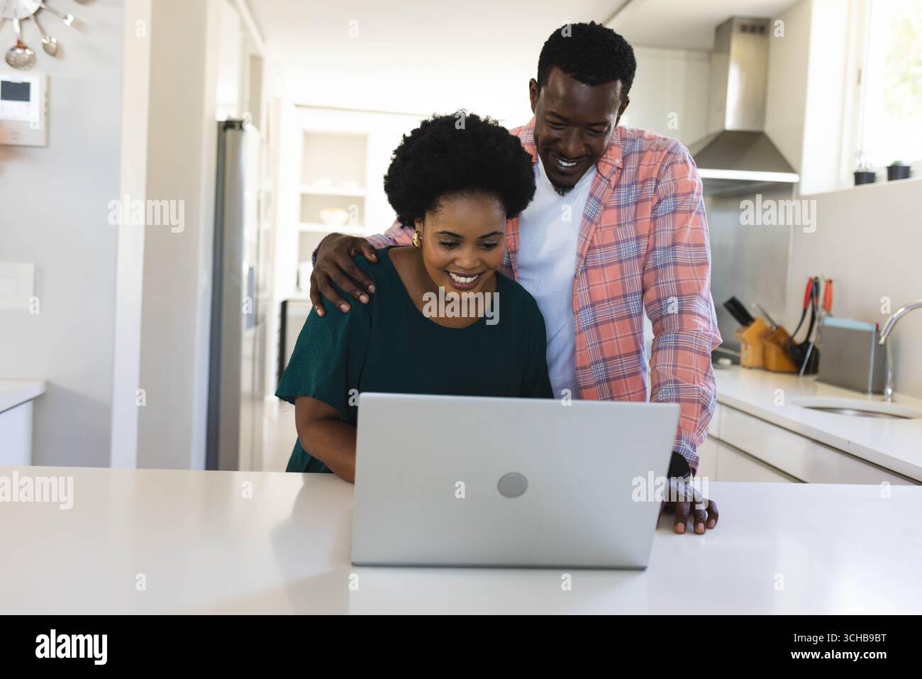 Coppia afroamericana pendente che ride mentre usa un laptop su un'isola di cucina, con pentole appese. Contemporaneo, stile di vita, minimalista, domestico, colla Foto Stock