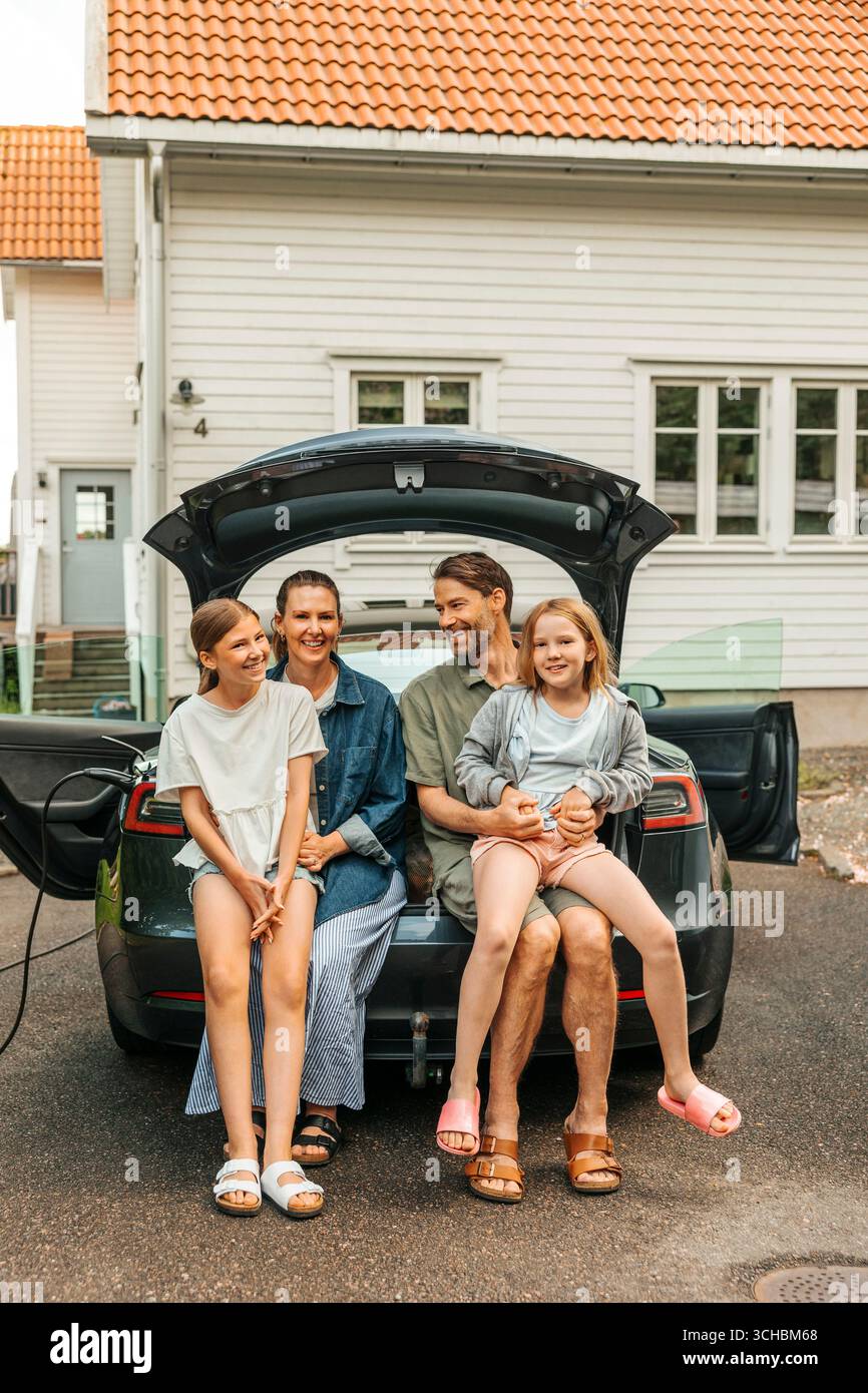 Uomo e donna sorridenti seduti con le figlie nel bagagliaio dell'auto elettrica sul vialetto Foto Stock
