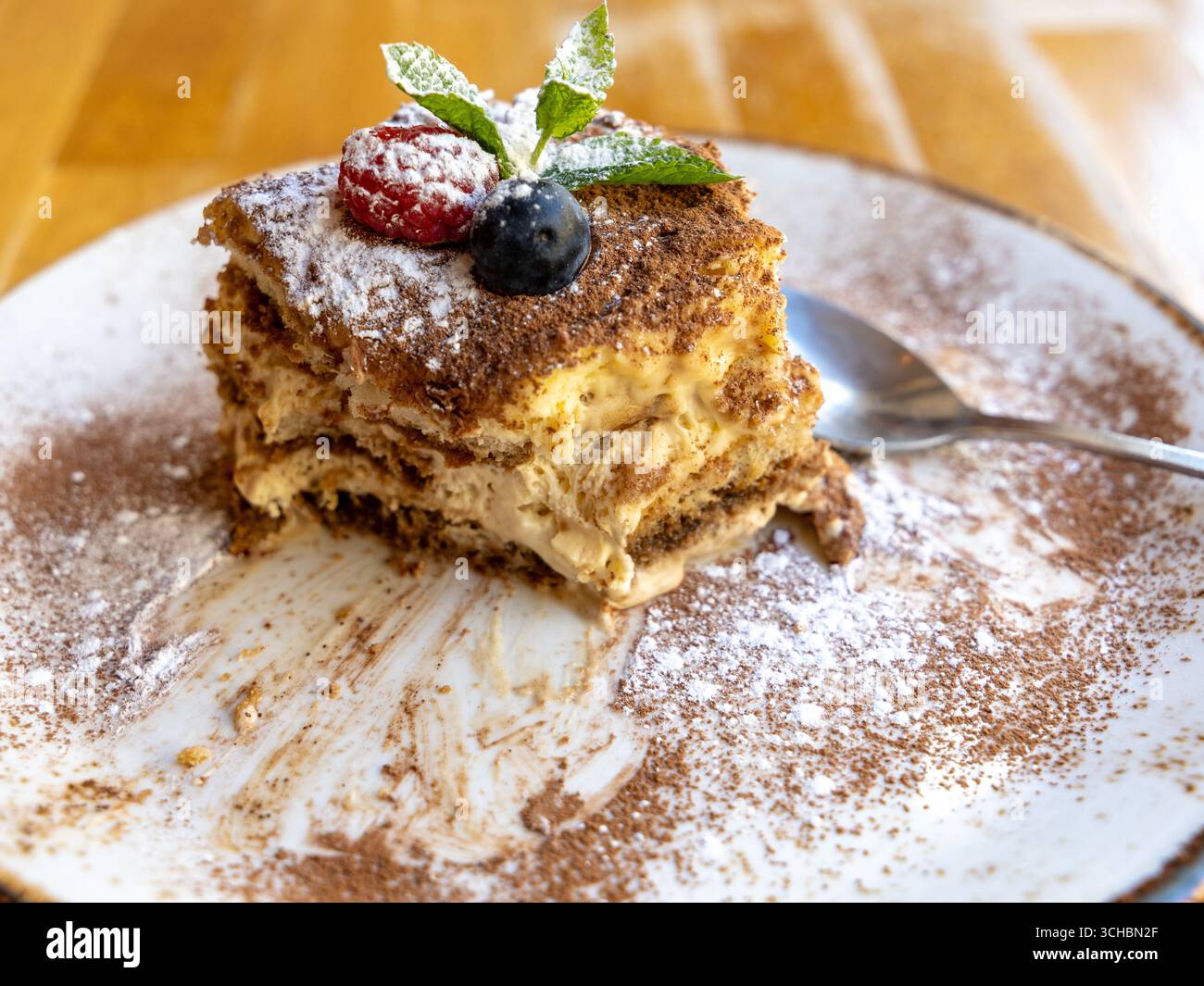 Primo piano di una torta tiramisù parzialmente mangiata con frutti di bosco e foglie di menta su un piatto spolverato di cannella con un cucchiaio Foto Stock