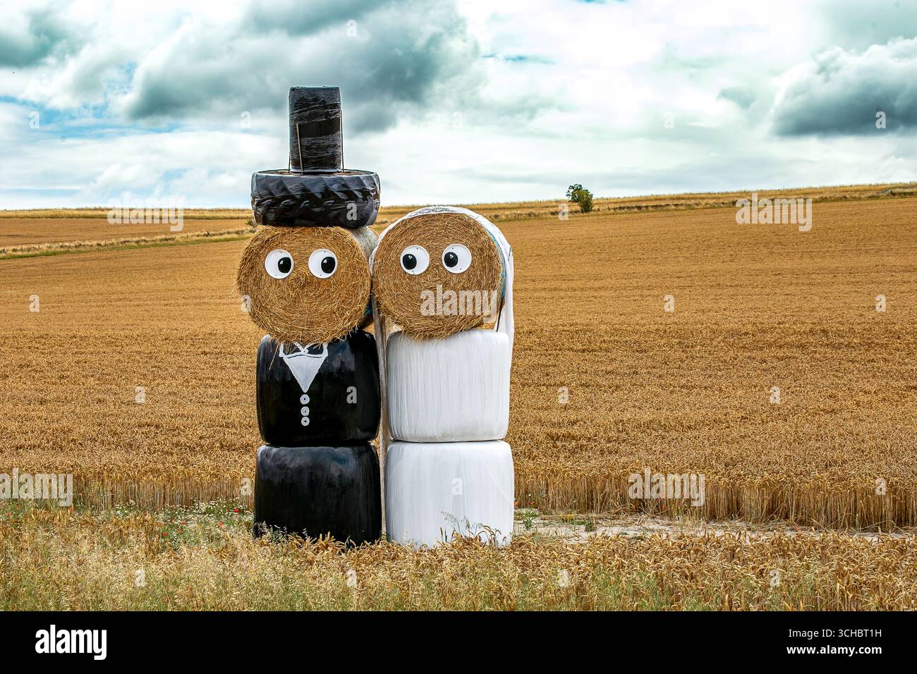 Balle di fieno decorate come una sposa e uno sposo in piedi in un campo di grano sotto un cielo nuvoloso che simboleggia la celebrazione del matrimonio rurale Foto Stock