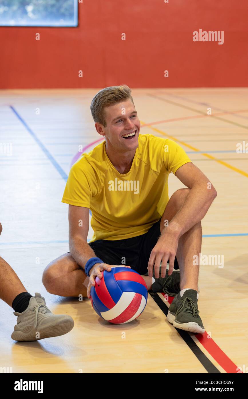 Uomo seduto sul campo da ginnastica con pallavolo e indossa camicia e braccialetto gialli. Fitness, sport, atletismo, lavoro di squadra, attivo, ricreazione, guarigione Foto Stock