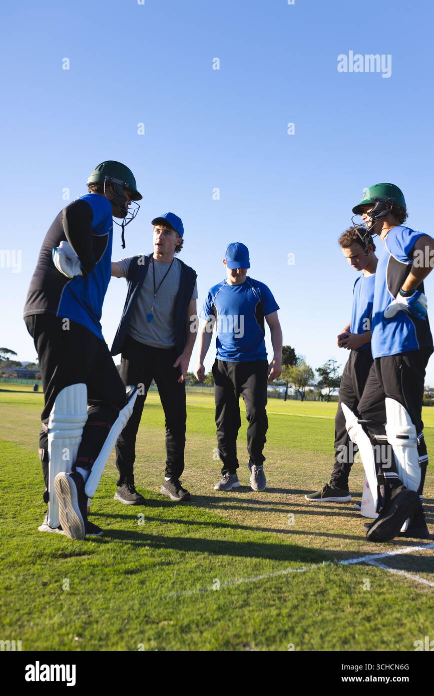 Squadre di cricket maschili diverse che si aggirano intorno al coach sul campo in erba indossando caschi e piazzole. Atletica, lavoro di squadra, strategia, outdoor, vibrante, competitivo, Foto Stock