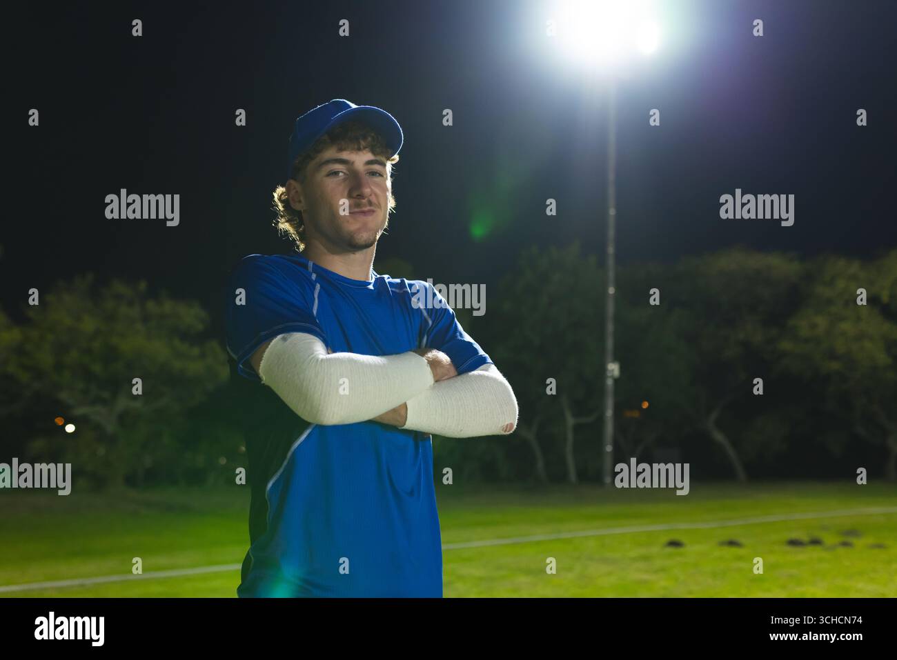 Giocatore di baseball maschile con cappello blu e maglia in piedi sul campo sportivo sotto il riflettore di notte. Atleta, sport, attività ricreative, all'aperto, di notte, Foto Stock