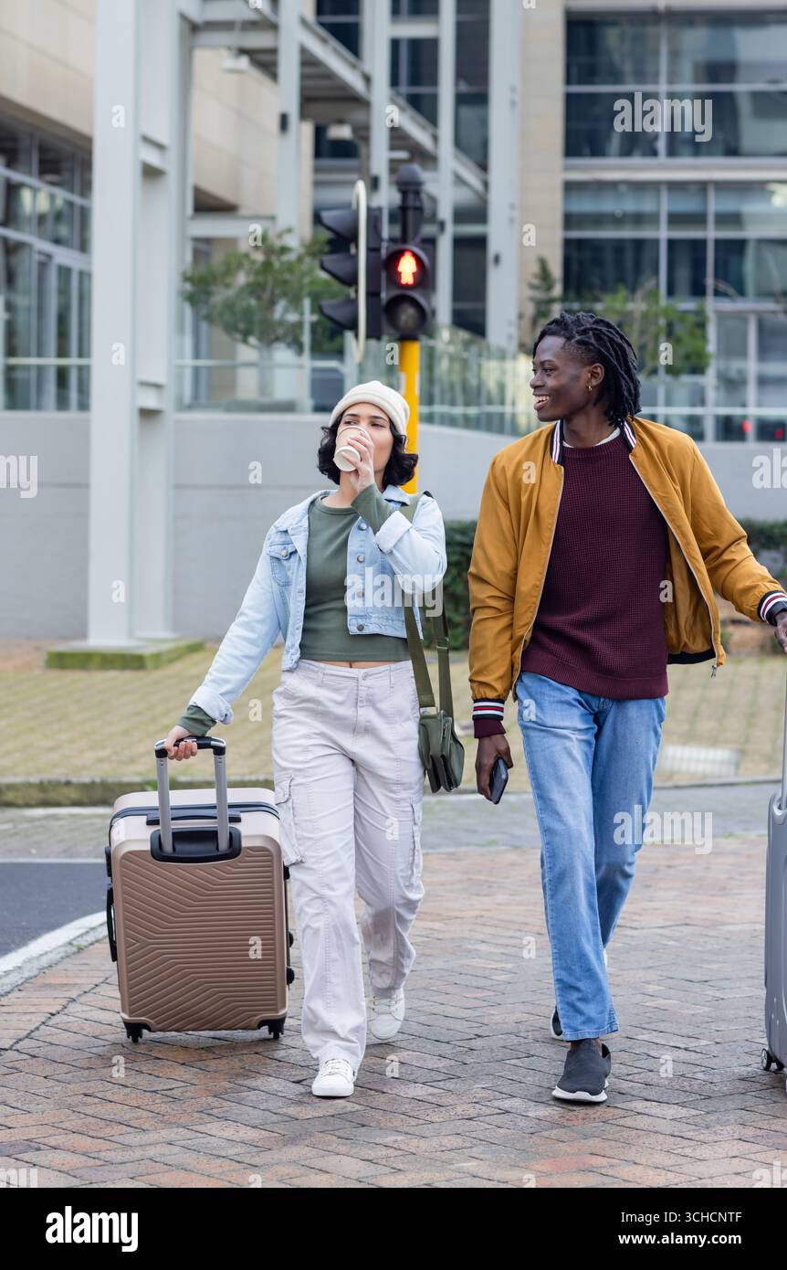 Diverse coppie che camminano dall'altra parte della strada tirando i bagagli e sorseggiando dalla tazza del caffè tramite il segnale incrociato. Urbano, viaggi, moderno, stile di vita, informale, transpo Foto Stock