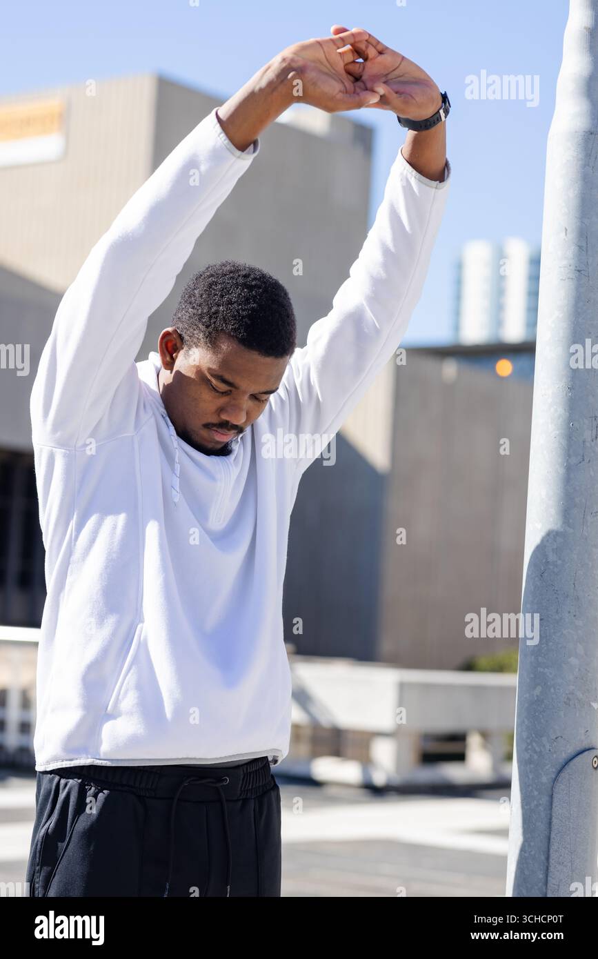 Uomo afro-americano che allunga le braccia sopra il tetto in pantaloni neri con cappuccio e orologio da polso bianchi. Urbano, fitness, stile di vita, moderno, attivo, all'esterno Foto Stock