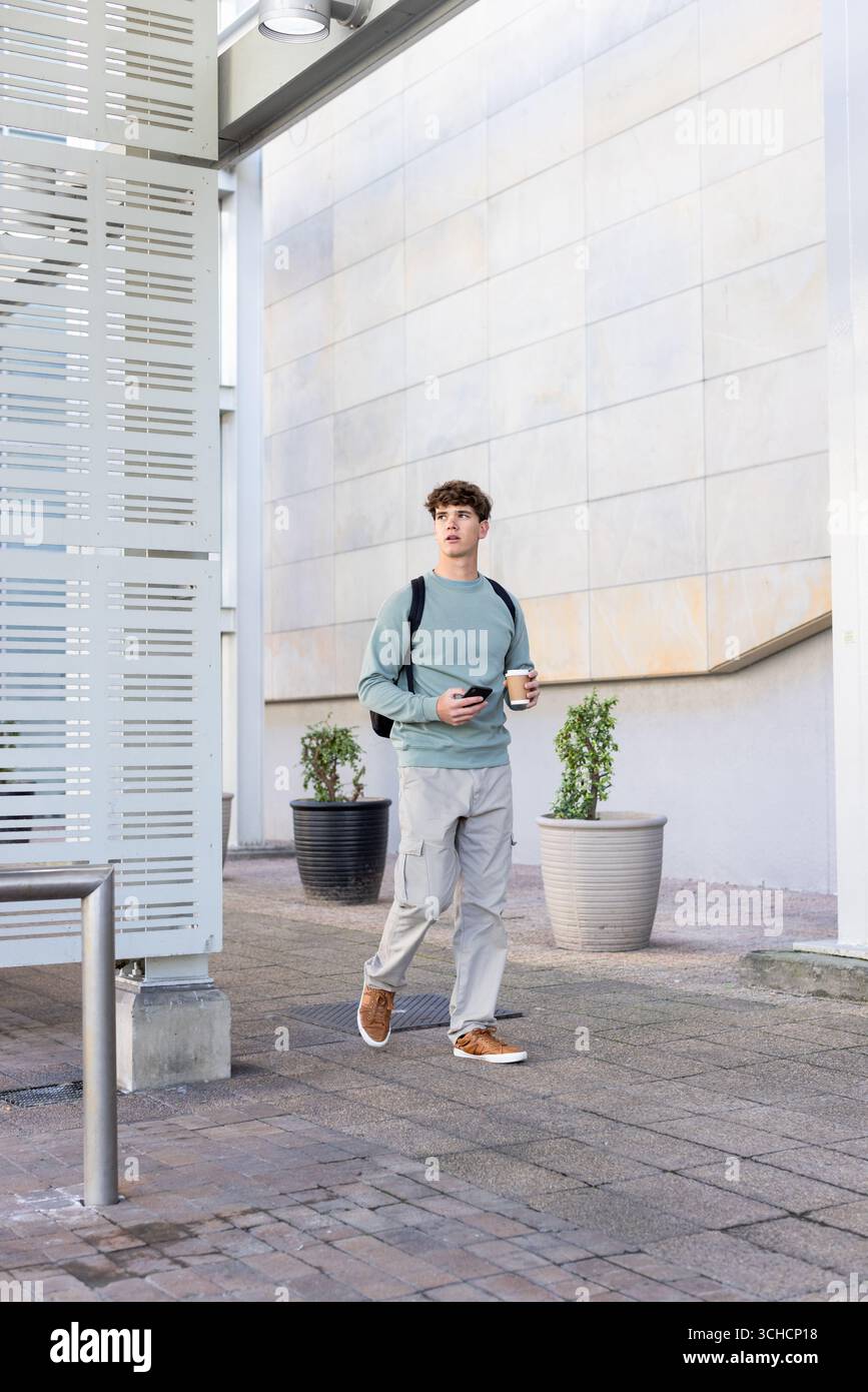 Uomo che cammina lungo la piazza lastricata accanto a un edificio moderno che porta con sé una tazza di caffè, uno smartphone e uno zaino. Urbano, moderno, contemporaneo, minimalismo, professione Foto Stock
