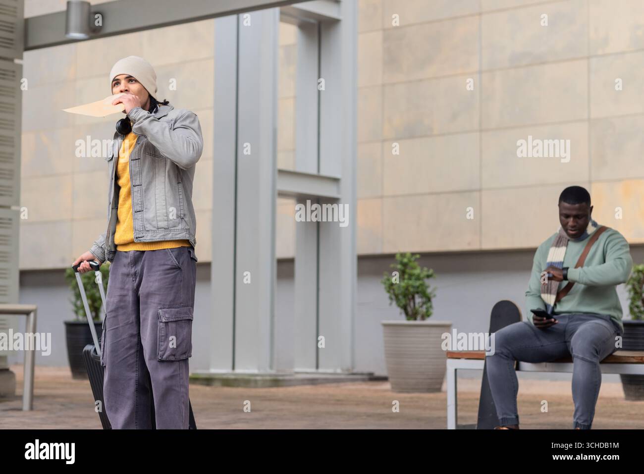Due uomini fuori dall'edificio che tengono la busta e tirano la valigia, controllano l'orologio e il telefono Foto Stock