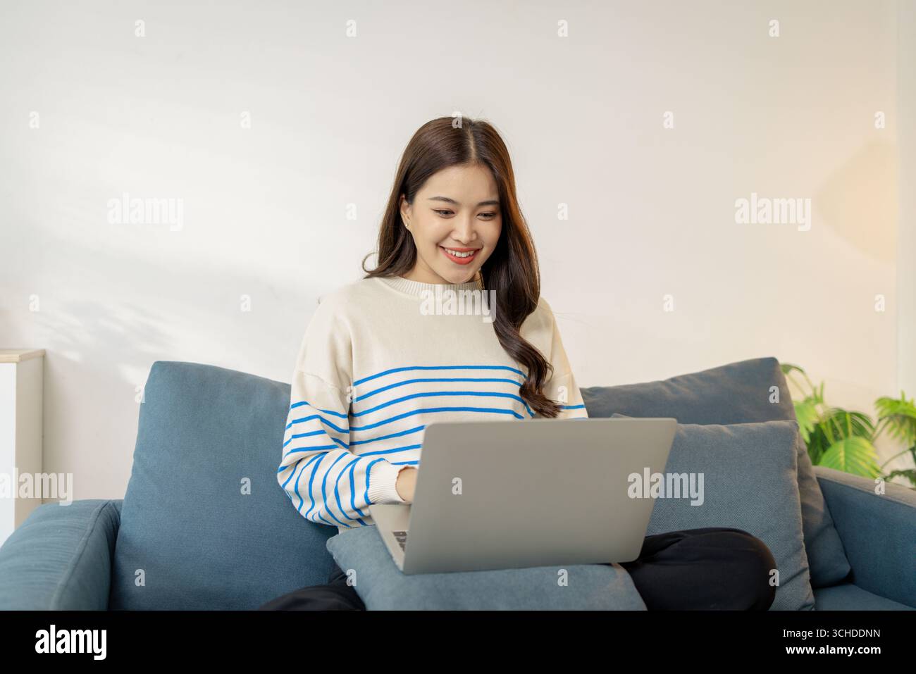 Una giovane donna è seduta comodamente su un divano utilizzando un computer portatile, sorridendo con concentrazione e gioia. Sembra impegnata nel suo lavoro o nella comunicazione online, crea Foto Stock