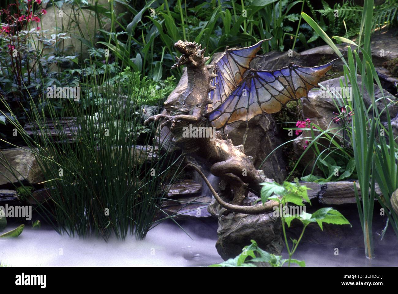 Una scultura di drago con ali colorate è posizionata tra piante vivaci e una nebbia delicata in un tranquillo giardino, creando un ambiente mistico. Foto Stock