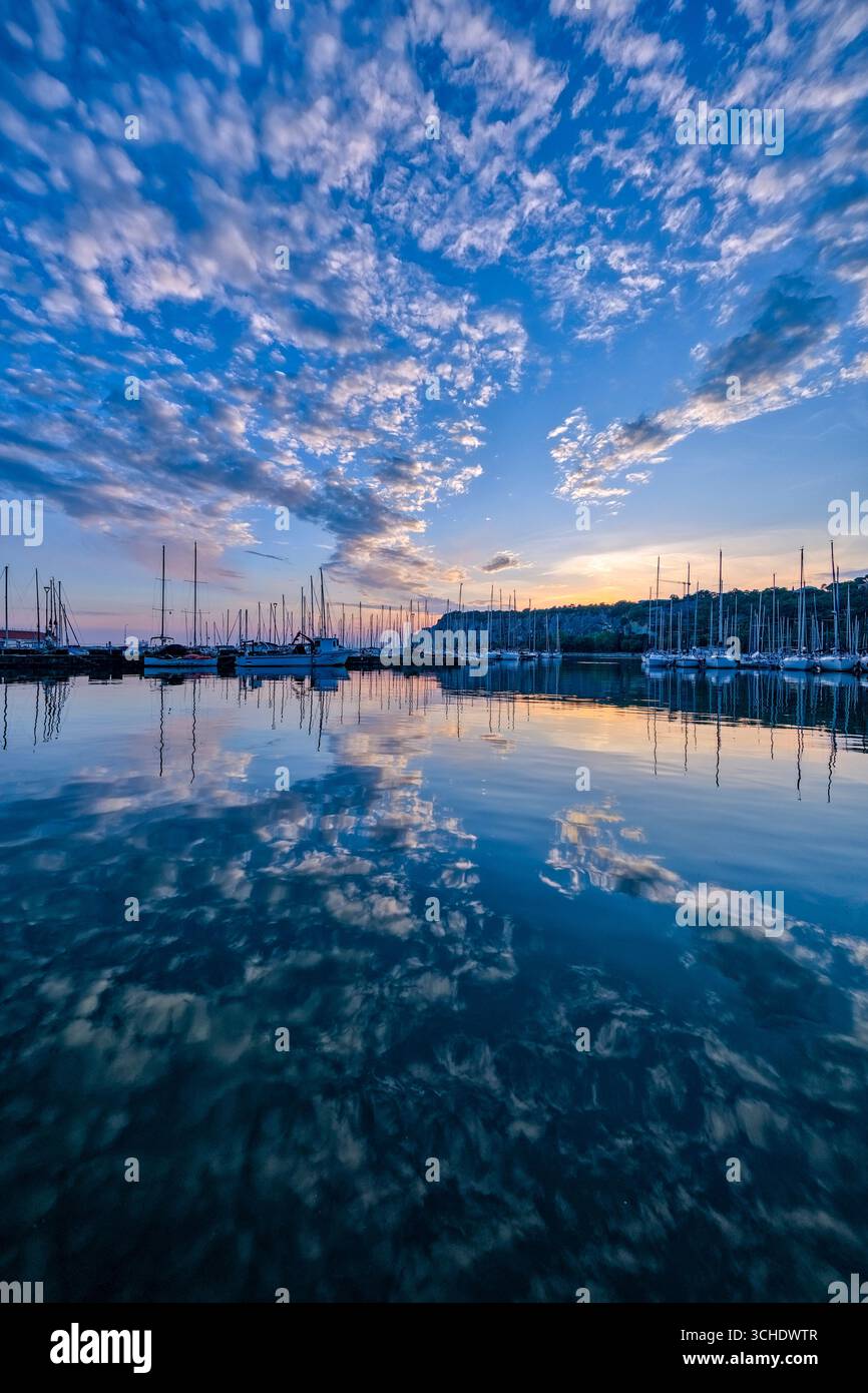 Il sole al tramonto, le nuvole colorate e le barche a vela si riflettono nelle acque del porto turistico di Castelreggio. Foto Stock
