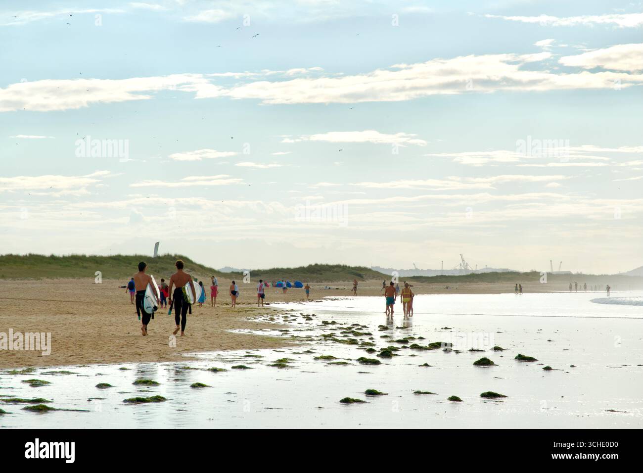Due giovani uomini che trasportano tavole da surf e turisti in una serata estiva sulla spiaggia di Somo famosa per il surf Ribamontan al Mar Cantabria Spagna Europa Foto Stock