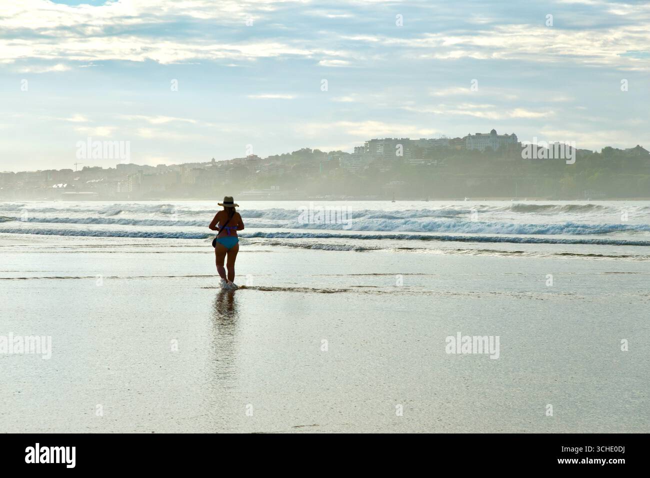 Donna solitaria che cammina in acque poco profonde sulla spiaggia di Somo famosa per il surf con la città di Santander e l'Hotel Real Ribamontan al Mar Cantabria Spagna Foto Stock