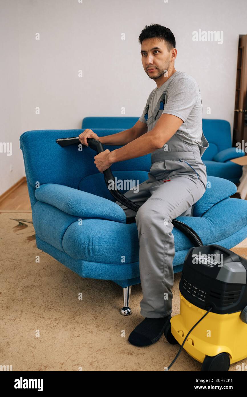 Ritratto verticale a tutta lunghezza del dipendente del servizio di pulizia professionale che indossa un'uniforme utilizzando un aspirapolvere per pulire il divano blu in soggiorno Foto Stock