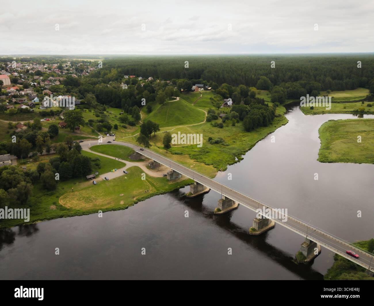 Vista aerea del ponte che si estende attraverso il fiume scuro verso il tumulo verde, con la foresta oltre, Merkine, Lituania. Foto Stock