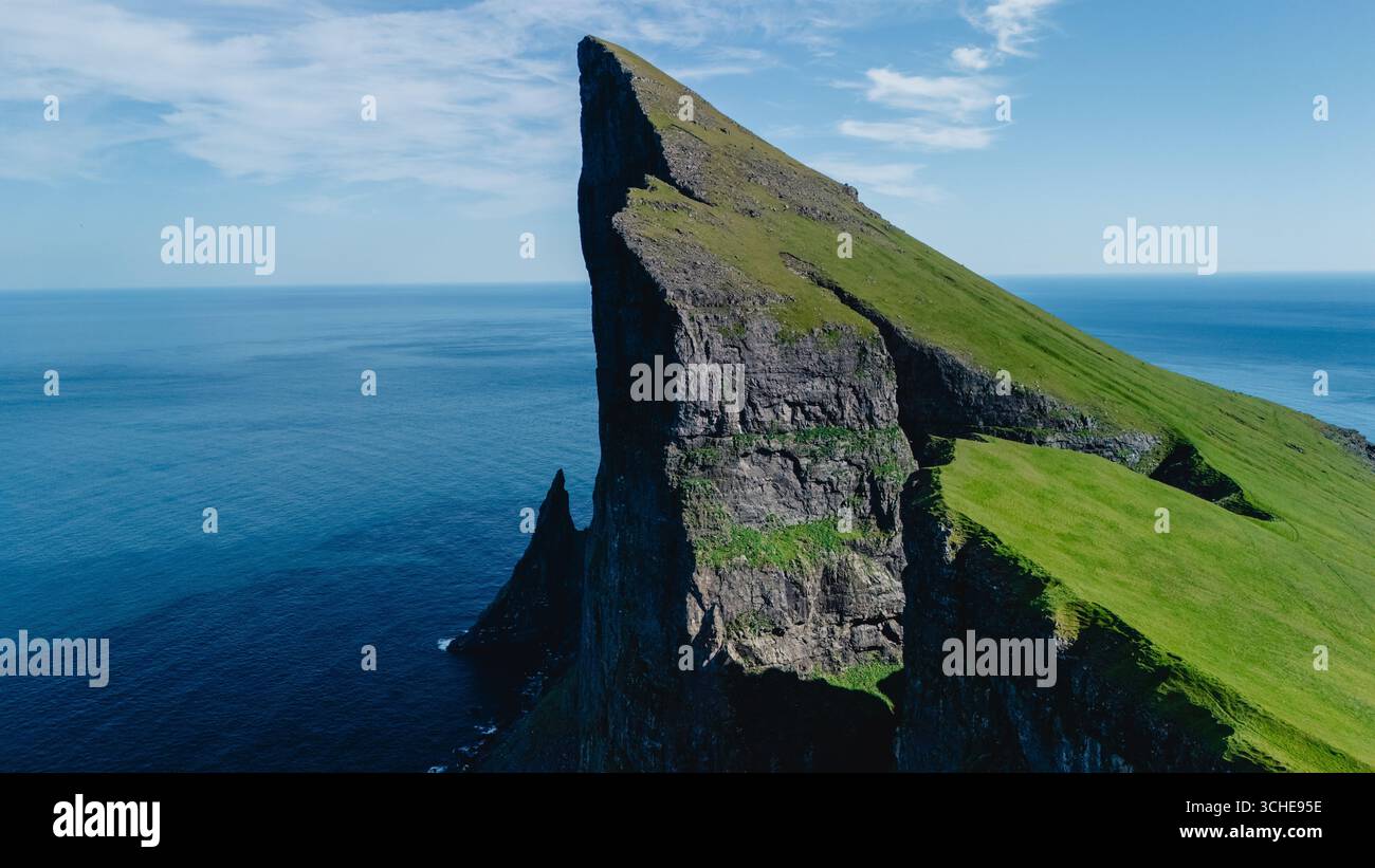 Una spettacolare scogliera sorge bruscamente dalle vibranti acque blu dell'Oceano Atlantico nelle Isole Faroe. La lussureggiante vegetazione contrasta con i bordi rocciosi, creando un panorama naturale mozzafiato. Foto Stock