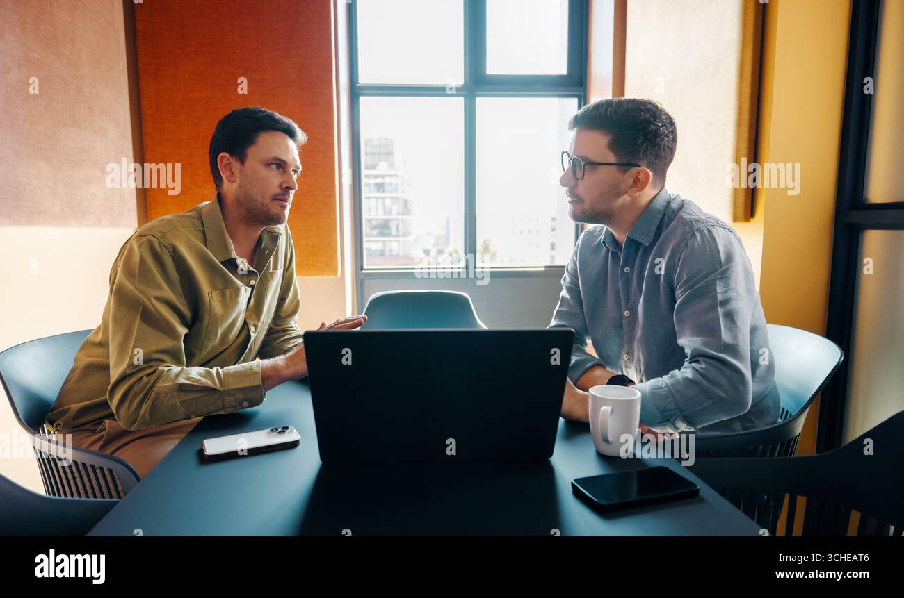 Due giovani adulti si impegnano in una discussione di lavoro mirata in un'atmosfera professionale e luminosa. Un laptop e uno smartphone sul tavolo suggeriscono Foto Stock