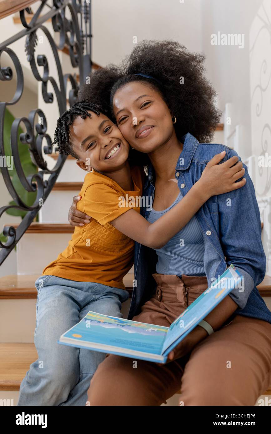 Madre e figlio seduti sulle scale, abbracciati e sorridenti con un libro aperto Foto Stock