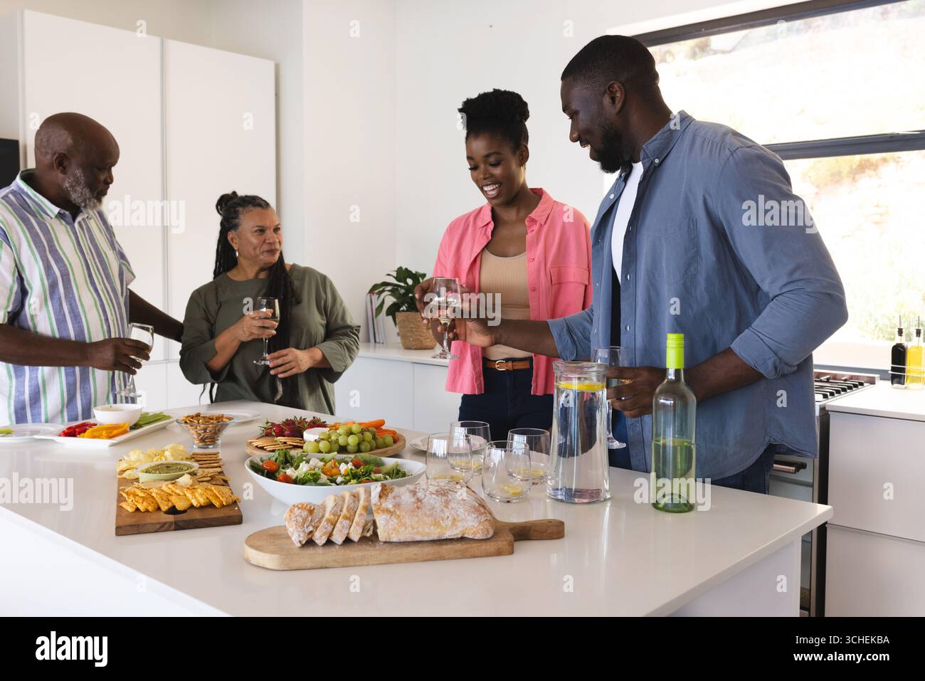 La famiglia si diverte con drink e stuzzichini in cucina moderna, condividendo risate Foto Stock
