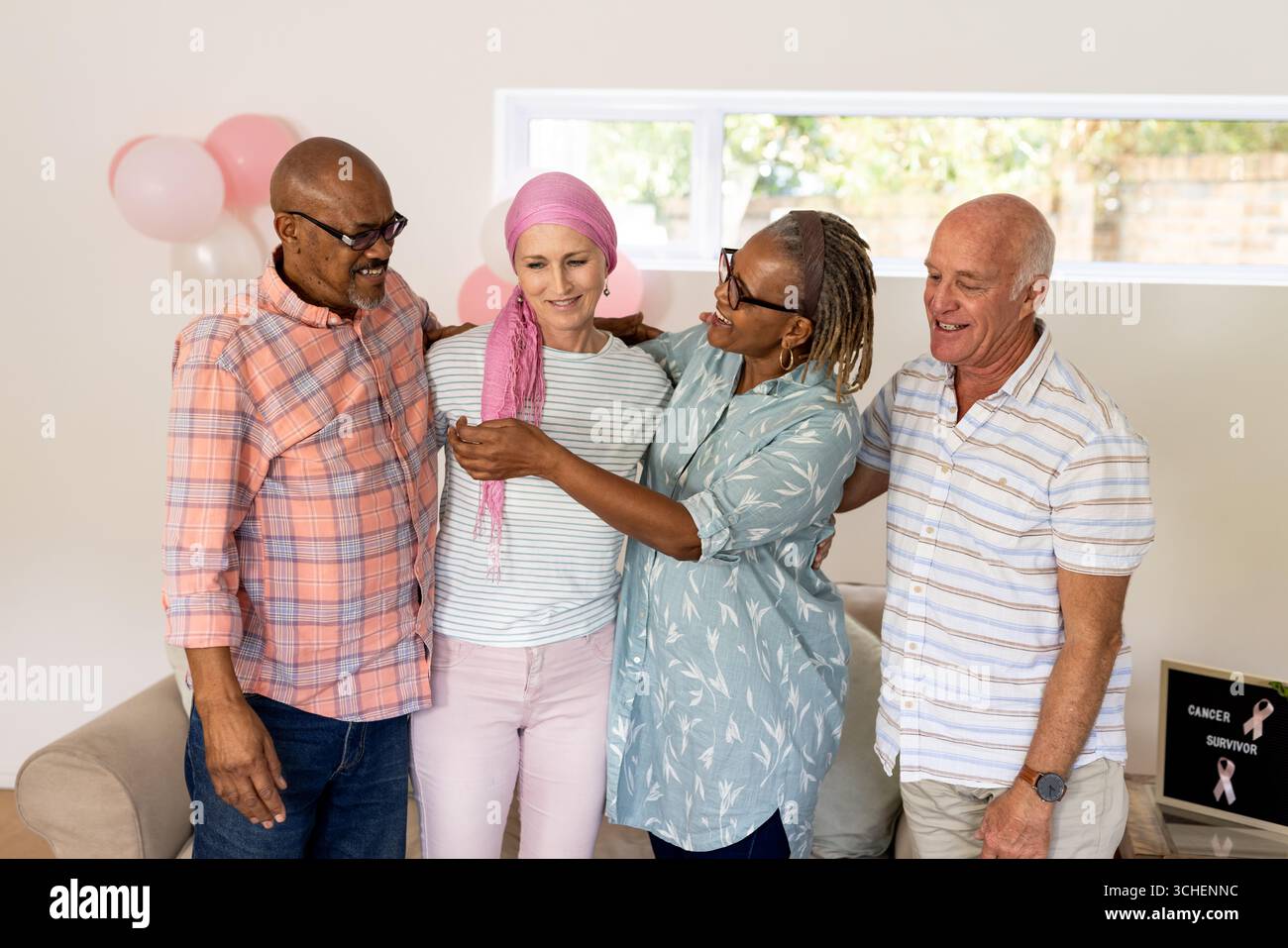 Amici anziani e diversi che sostengono la sopravvissuta al cancro con il nastro rosa a casa, sorridendo insieme Foto Stock