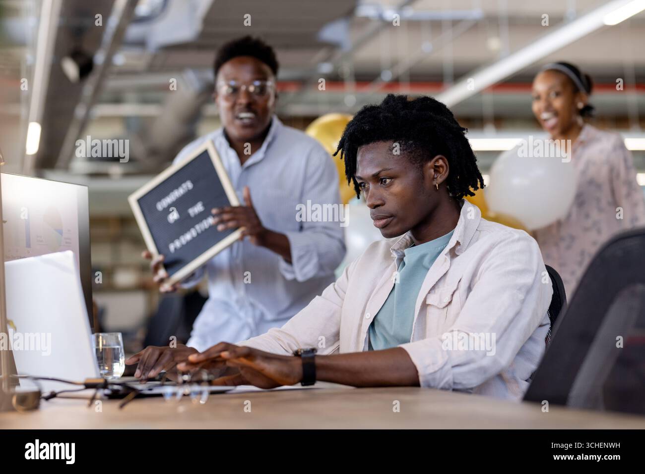 Un giovane che scrive sul laptop in ufficio mentre colleghi diversi festeggiano nelle vicinanze Foto Stock