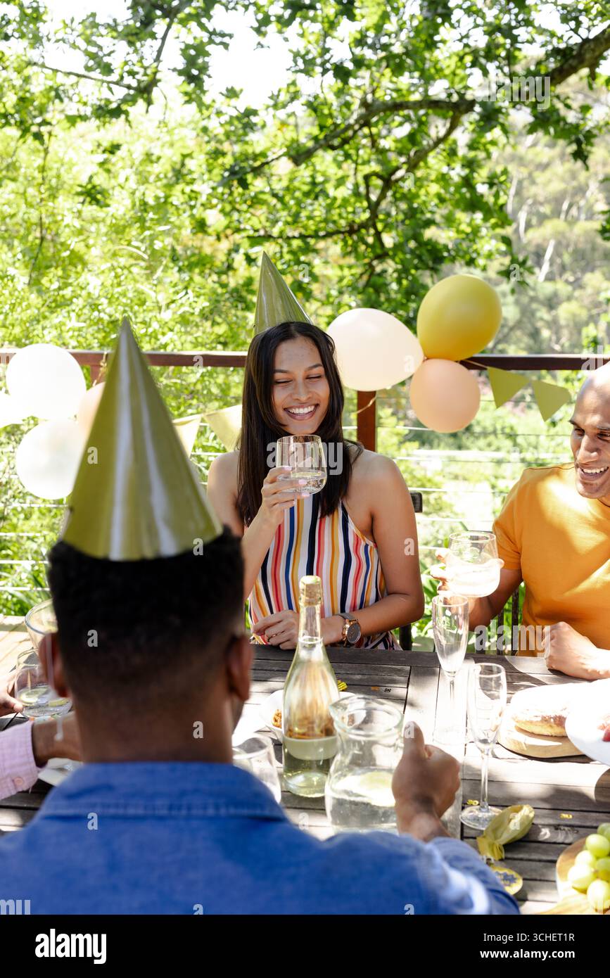 Festeggiate con bevande, amici diversi che indossano cappelli da festa e condividono risate all'aperto in giardino Foto Stock