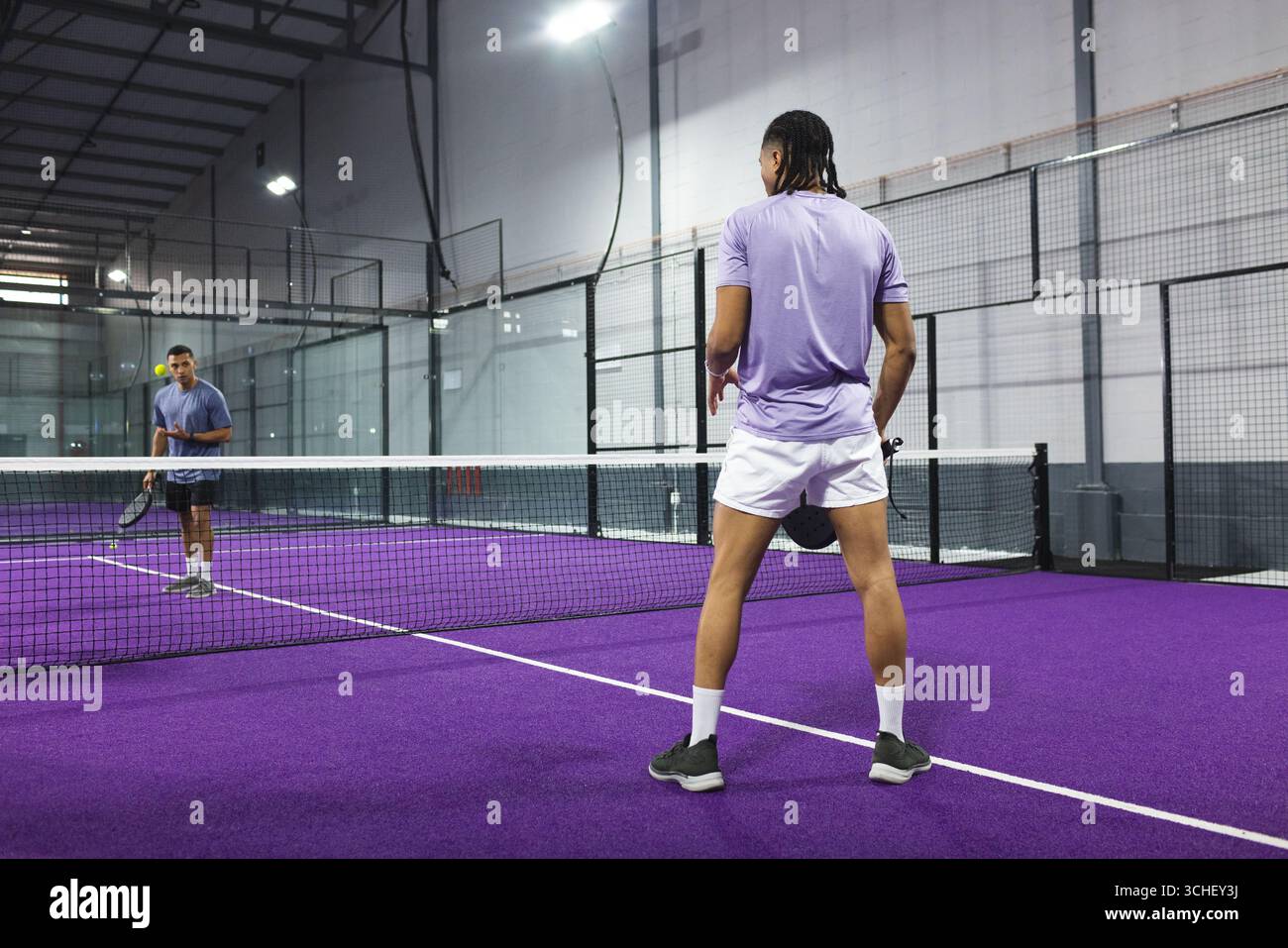 Diversi avversari maschili che giocano su un campo di padel viola con rete, racchette e palla gialla, spazio copia Foto Stock
