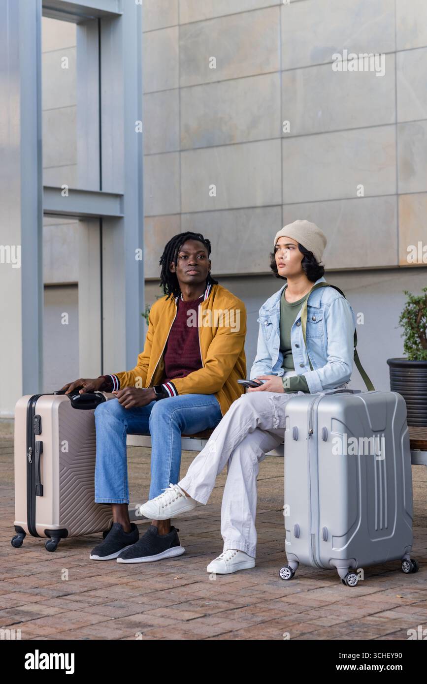 Diversi compagni di viaggio aspettano sulla panchina presso l'hub di transito, con valigie rotanti e smartphone Foto Stock Diversi compagni di viaggio aspettano sulla panchina presso l'hub di transito, con valigie rotanti e smartphone Foto Stock