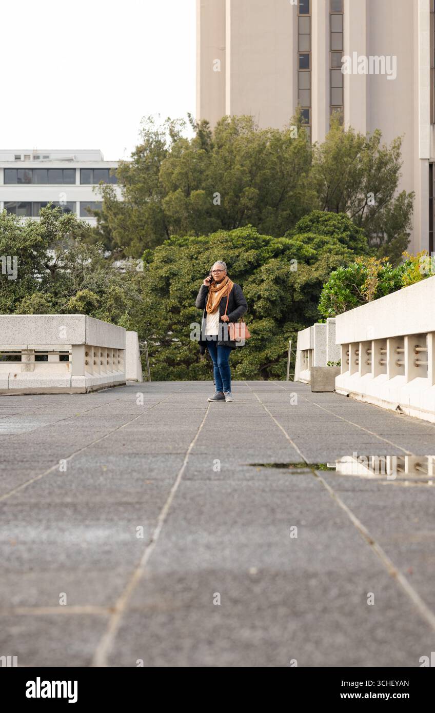 Donna afroamericana anziana sul ponte che parla al telefono con borsa abbronzante, ringhiere, pozzanghera Foto Stock