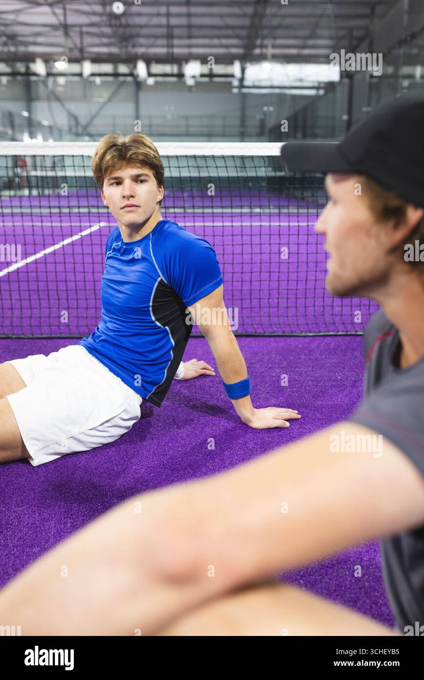 Compagni di squadra maschili che riposano su un campo viola accanto alla rete in una struttura sportiva con braccialetto e berretto Foto Stock