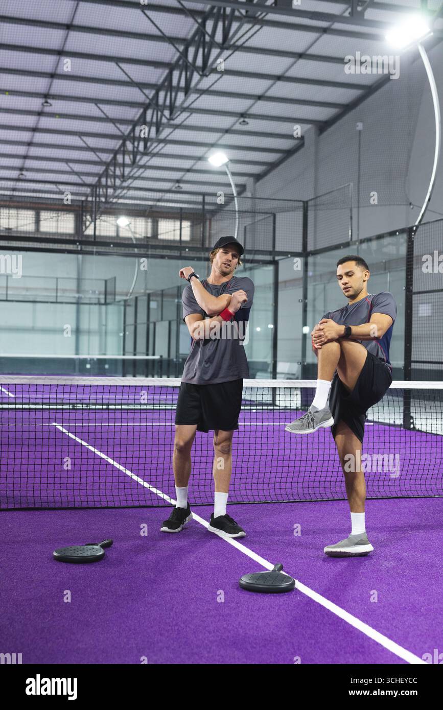 Due giocatori di padel maschili che si stendono su un campo di padel viola in una struttura con tetto in metallo con rete e racchette Foto Stock
