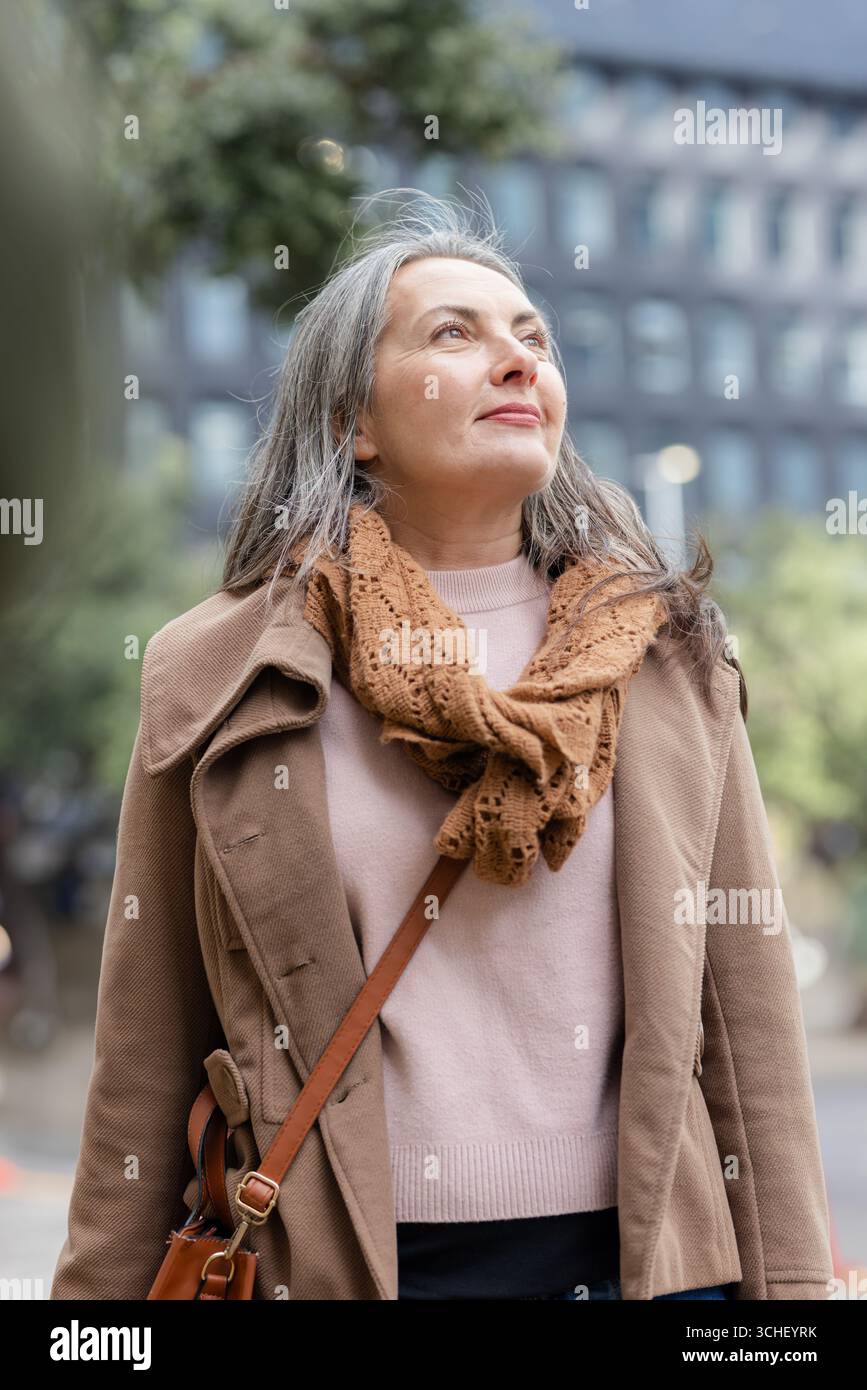 Donna senior con cappotto marrone e sciarpa a maglia marrone che trasporta una borsa a tracolla in pelle in Urban plaza Foto Stock