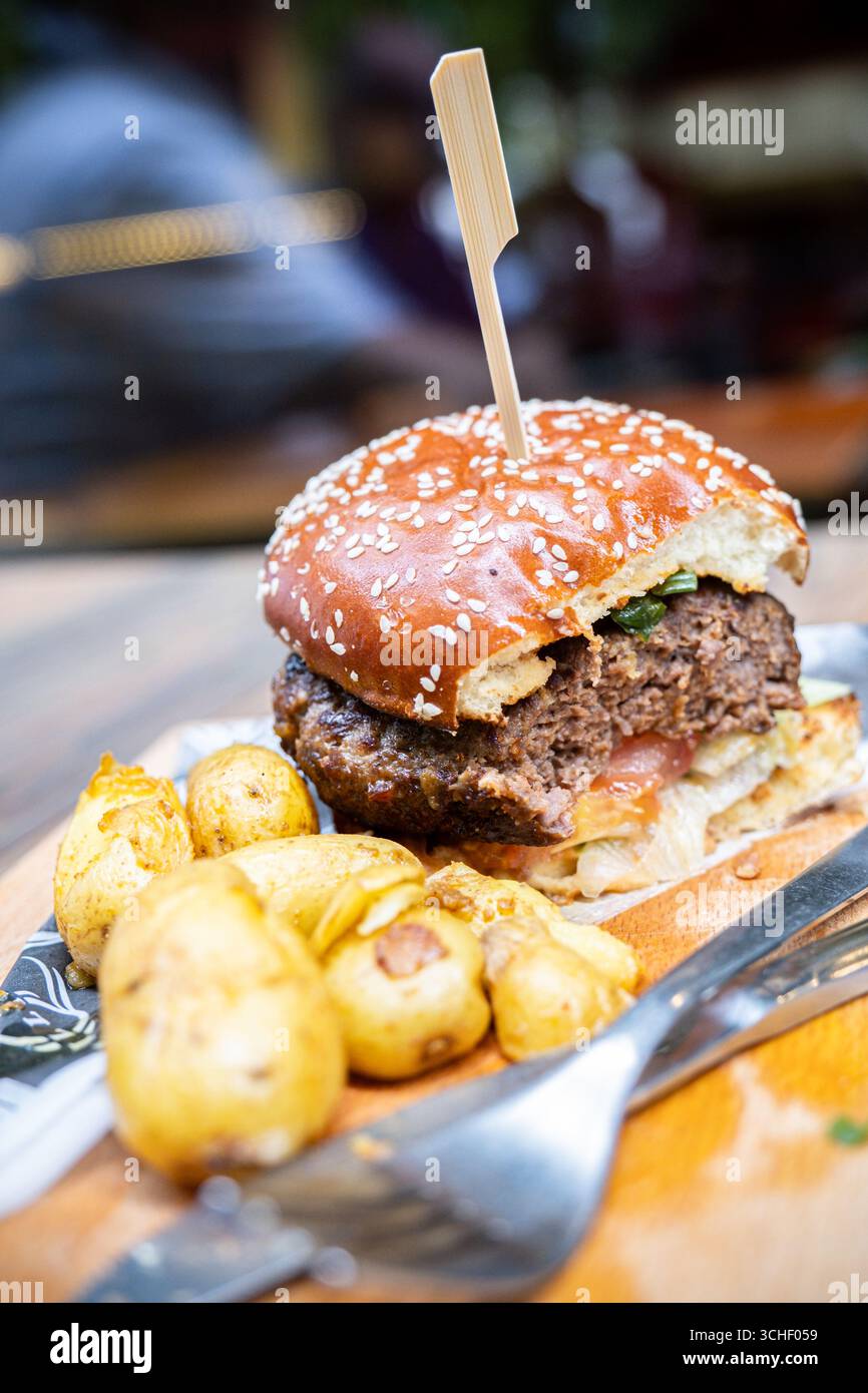 Hamburger succoso con panino di semi di sesamo, guarnito con verdure fresche, servito insieme a croccanti patate arrosto su un piatto di legno Foto Stock