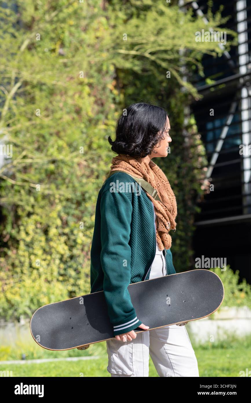 Donna asiatica che cammina accanto all'edificio rivestito di edera tenendo lo skateboard sull'erba con la sciarpa Foto Stock