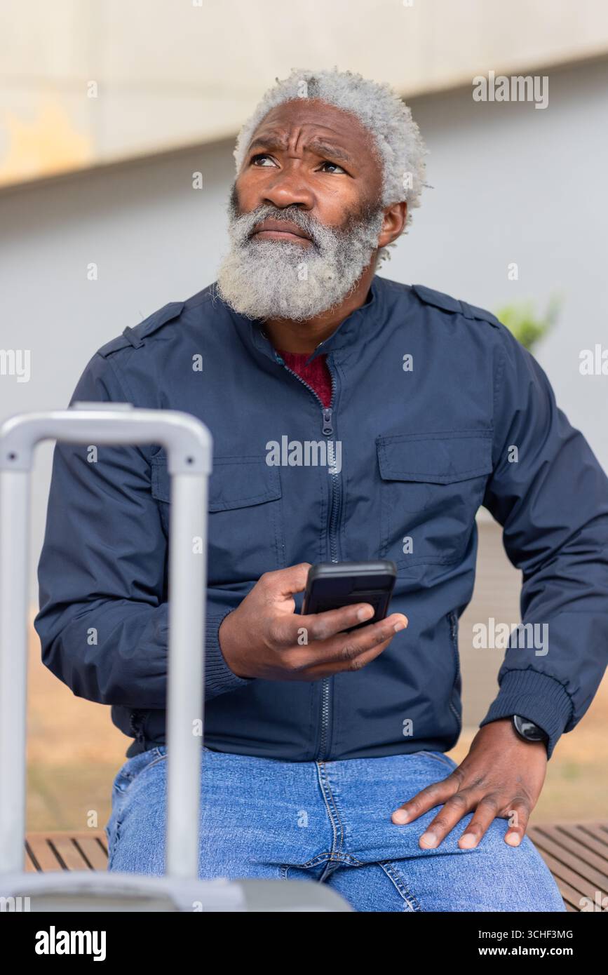 Uomo afroamericano anziano in giacca seduto sulla panchina al plaza con lo smartphone con la valigia Foto Stock
