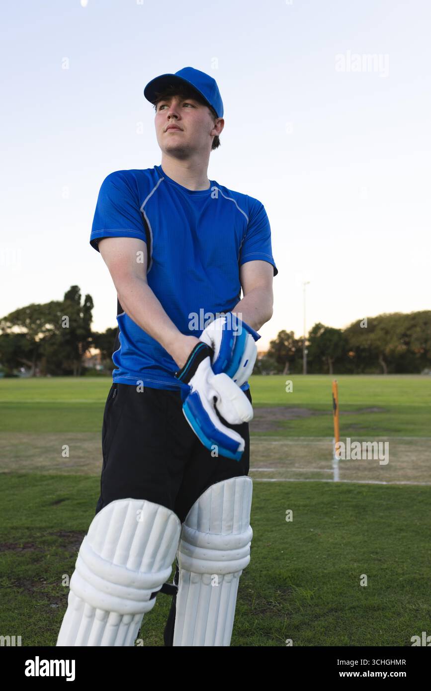 Giocatore di cricket maschio che tira i guanti da battuta e fissa i cuscinetti delle gambe sul campo di cricket con il cappello blu Foto Stock