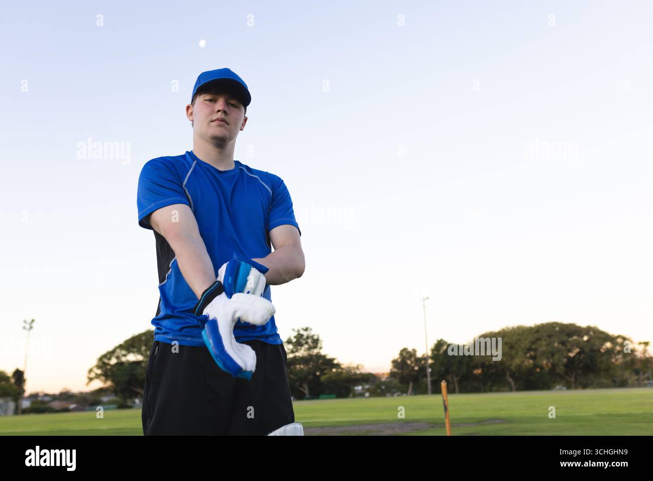 Cricketer maschio che tira i guanti da wicket-keeping da ceppi sul campo di cricket in maglia blu, berretto Foto Stock