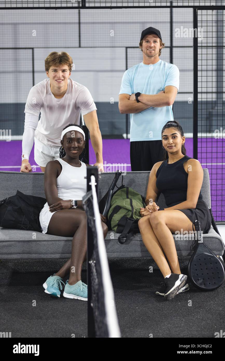 Amici diversi seduti e in piedi intorno alla panchina sul campo del padel con racchette e borsa sportiva Foto Stock