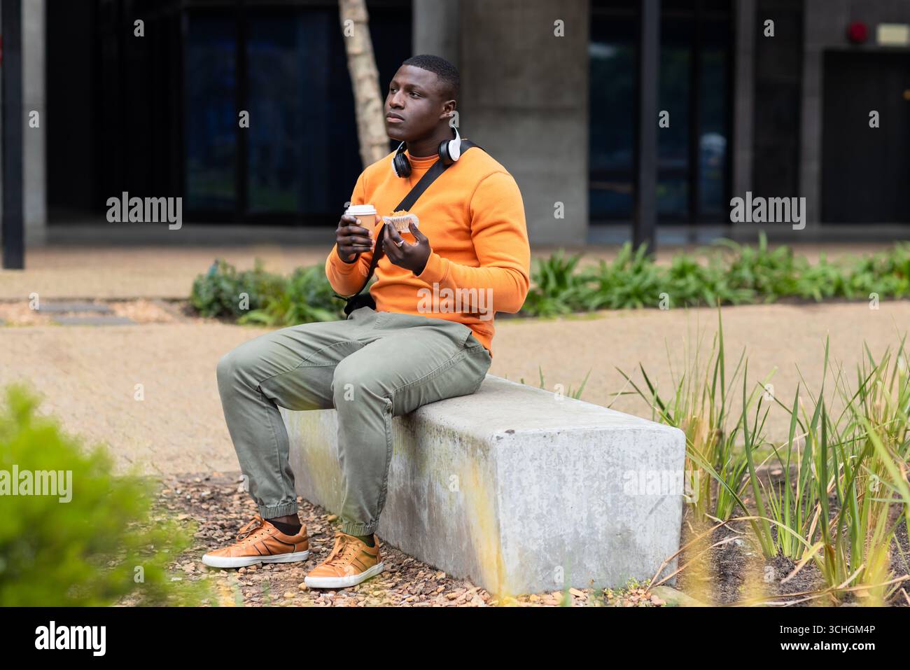 Uomo afroamericano seduto sulla panchina al plaza, con le cuffie e con caffè e pasticcini Foto Stock