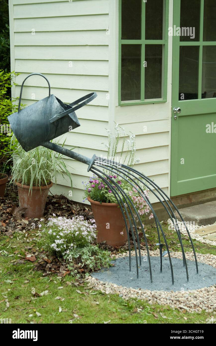 Una scultura in metallo creativa raffigurante una barattola che versa acqua e un rastrello, posizionata all'esterno di un capannone di giardino circondato da piante. Foto Stock