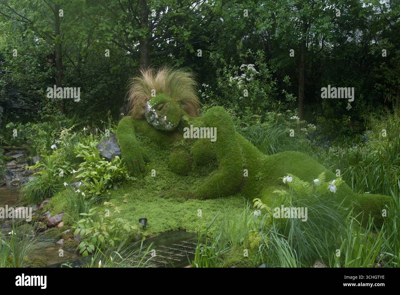 Una grande scultura ricoperta di muschio di una figura reclinabile si fonde con piante vibranti, mostrando l'arte della natura in un tranquillo giardino. Foto Stock