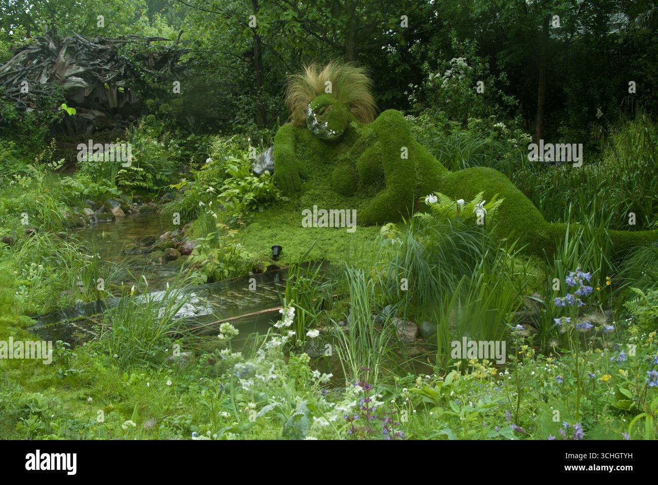 Una figura scultorea ricoperta di muschio giace pacificamente tra una vegetazione vibrante e fiori in fiore, creando una tranquilla oasi all'aperto. Foto Stock