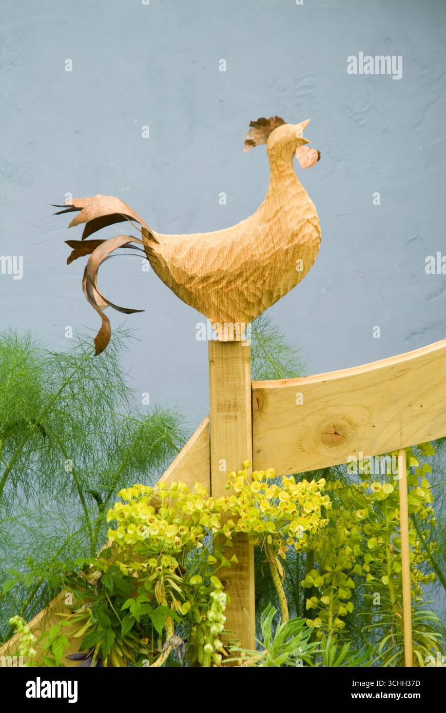 Una scultura in legno di gallo si erge orgogliosamente in cima a un palo circondato da lussureggianti piante verdi in un tranquillo ambiente esterno. Foto Stock
