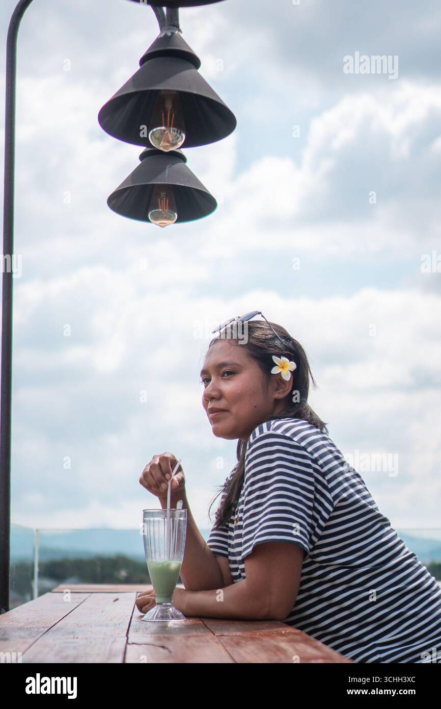 Una donna asiatica siede in un caffè sulla terrazza con una bevanda fresca, indossando camicia a righe e fiori sui capelli. Foto Stock