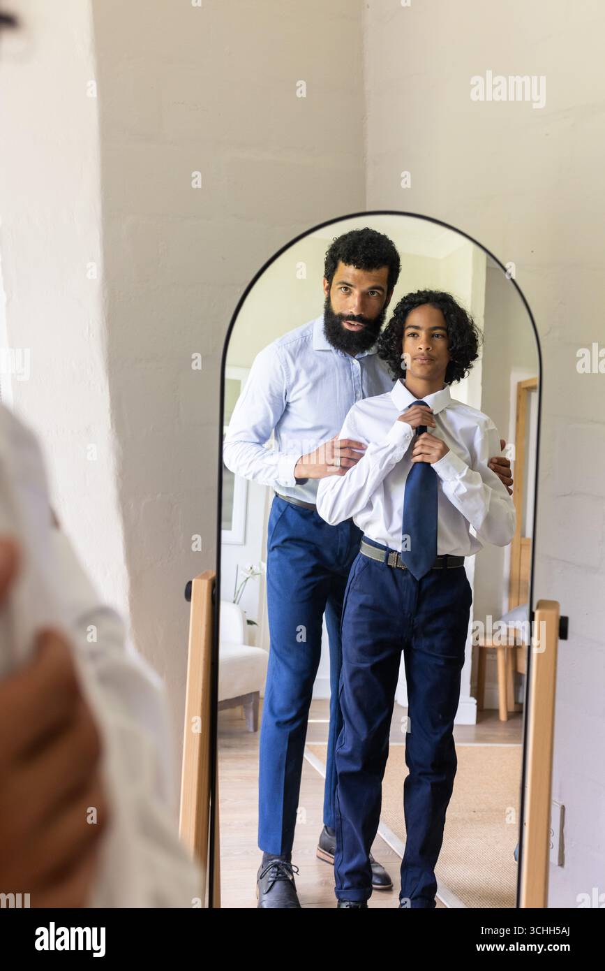 Famiglia variegata, padre aiuta il figlio a regolare la cravatta nello specchio, preparandosi per le occasioni speciali, a casa Foto Stock