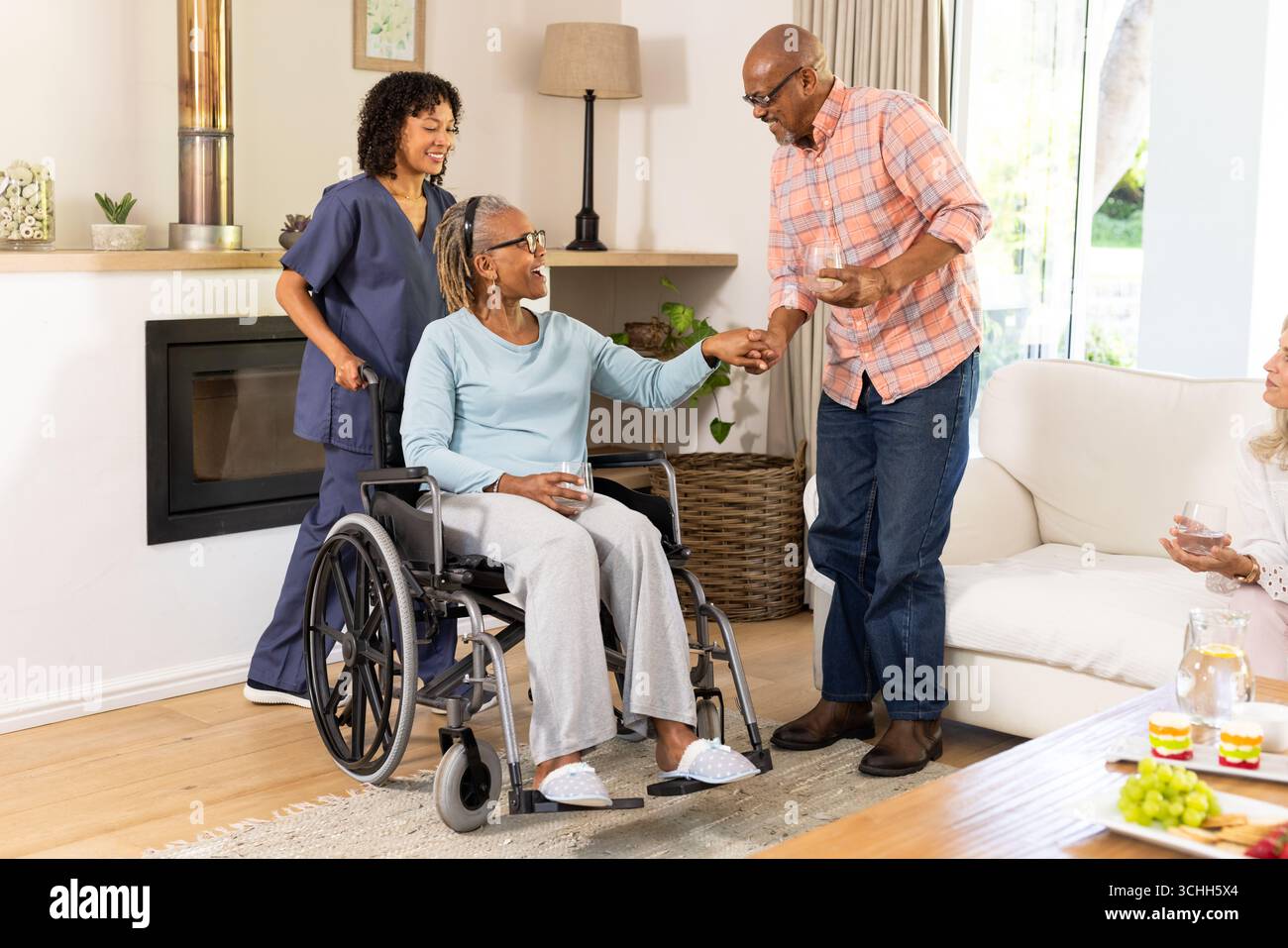 Uomo anziano che aiuta la donna in sedia a rotelle mentre l'assistente sorride a casa Foto Stock
