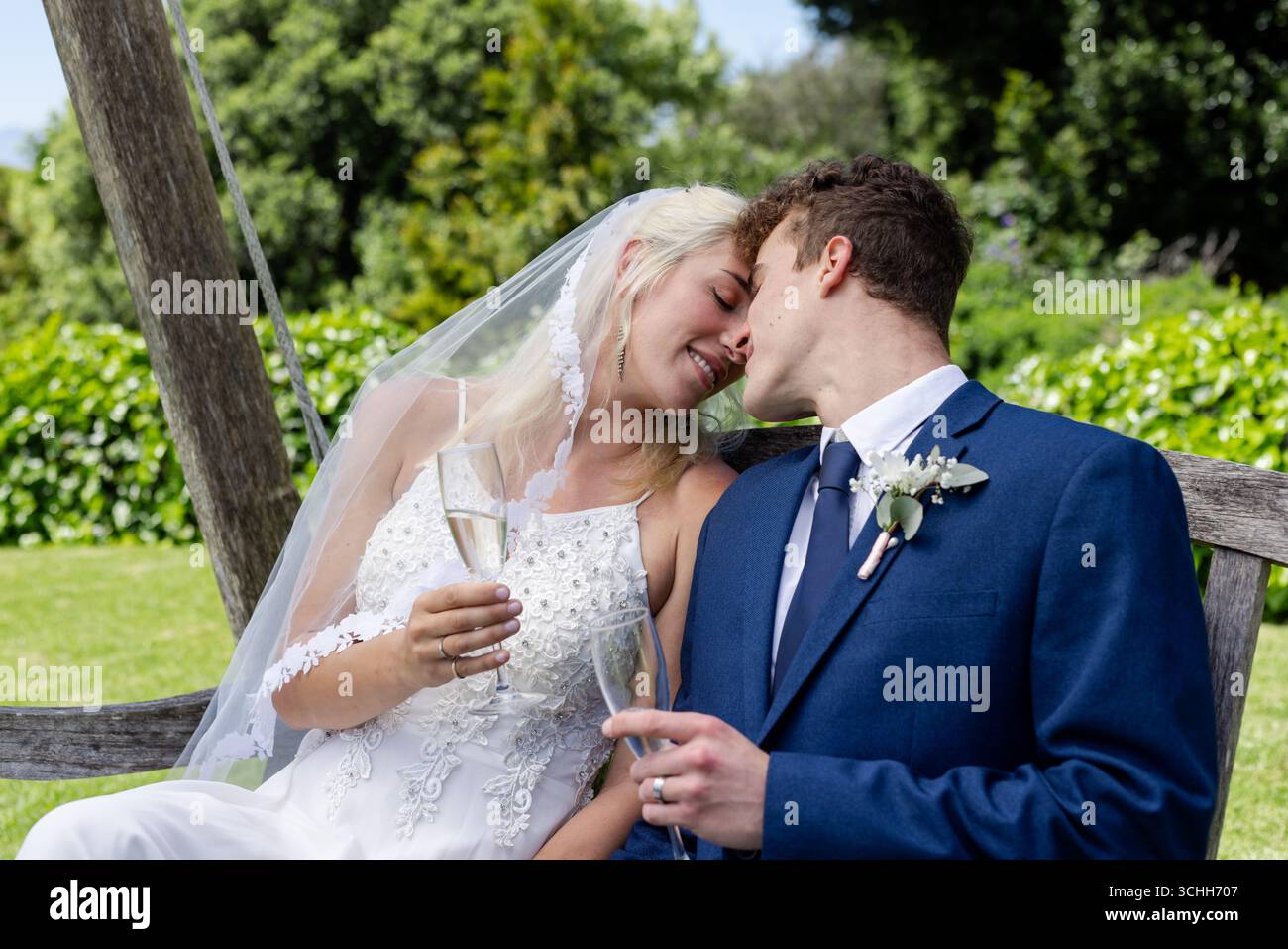Sposa e sposo condividono momenti romantici sulla panchina del giardino, tenendo in mano bicchieri da champagne Foto Stock