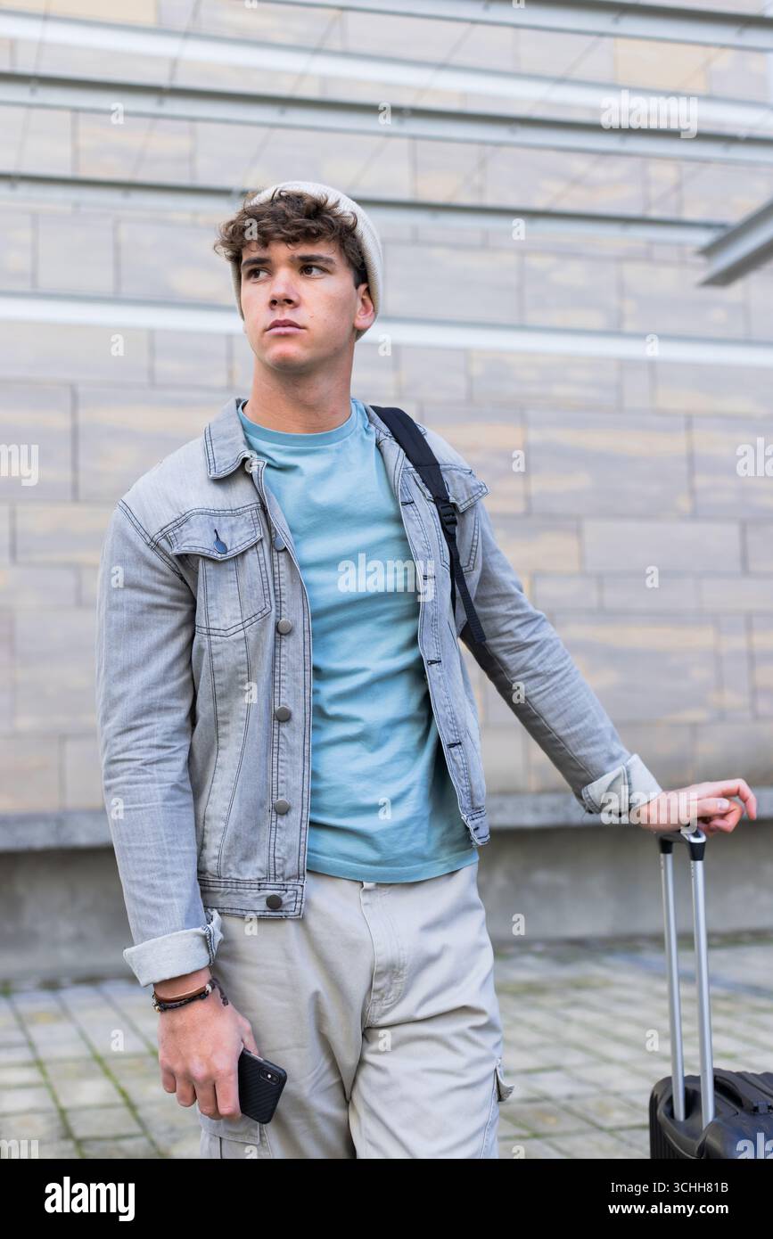 Uomo che tiene la maniglia della valigia e controlla lo smartphone sul vialetto pavimentato urbano accanto alla facciata Foto Stock