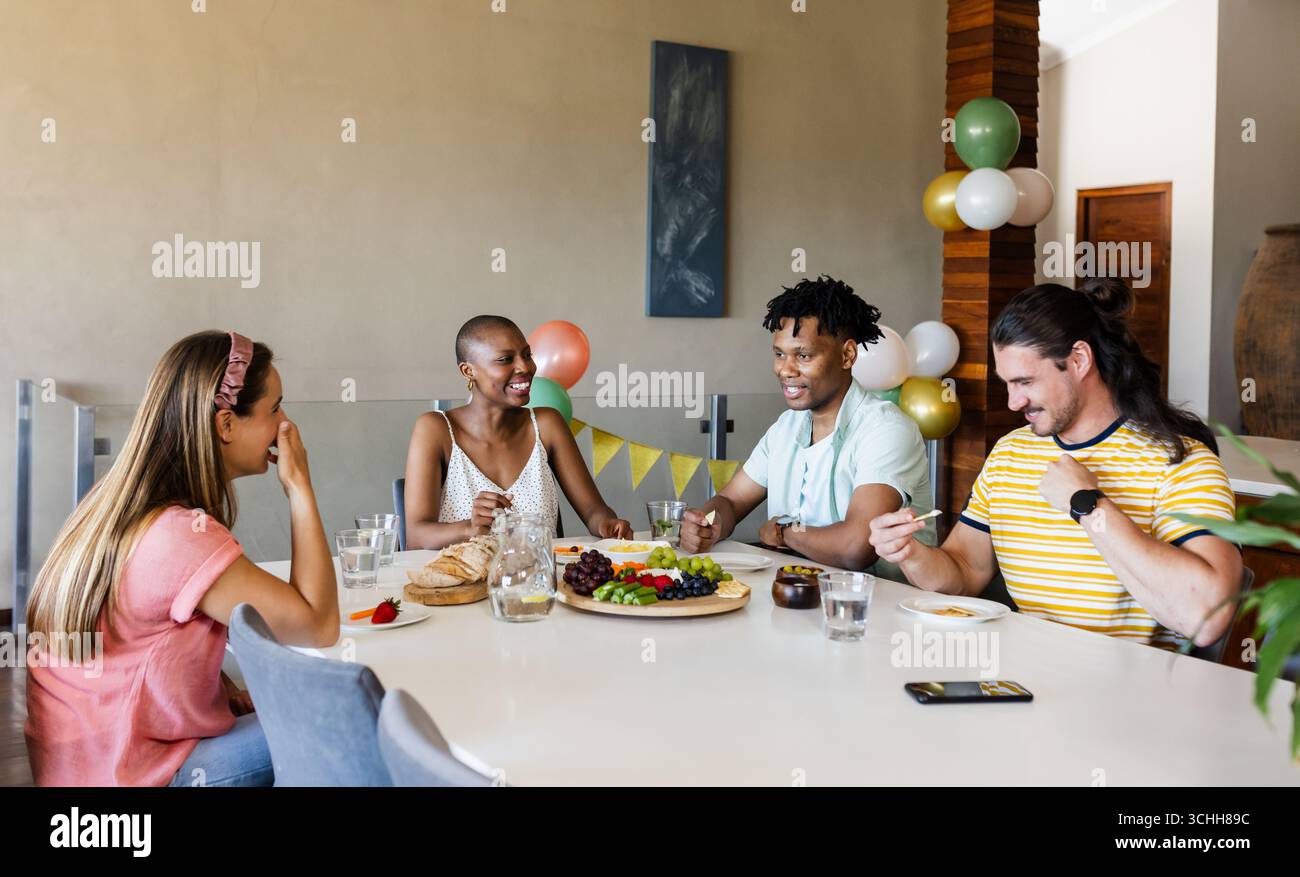 Amici diversi che mangiano insieme a casa, ridono e condividono storie Foto Stock